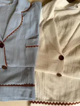 Pijama de algodón color blanco, compuesto por una camisa de manga corta abotonada con cuello solapa y un bolsillo en el pecho, y un short con elástico en la cintura. Ambas prendas tienen un detalle de picot marrón en los bordes.