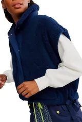 Chaleco de piel sintética tipo sherpa en color blanco crema, con cuello alto y cierre frontal mediante botones a presión. Presenta un diseño de hombros caídos y detalles de lazos decorativos en los laterales inferiores.