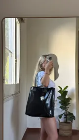 Bolso tote negro de cuero sintético con textura de cocodrilo. Presenta costuras decorativas en los laterales y un dije colgante en forma de cruz plateada con detalles de perlas.