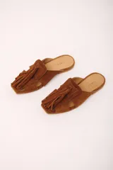 Zueco de cuero gamuzado color marrón, con punta cuadrada y detalle de gran borla de flecos en el empeine. Presenta aberturas laterales y suela plana.