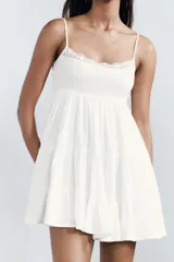 Vestido corto blanco de gasa con escote recto y tirantes finos ajustables. Presenta un detalle de encaje a tono en el borde del escote y forro interior.