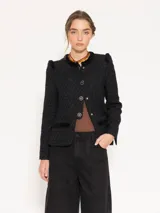 Chaqueta corta color negro confeccionada en jacquard chanel, con detalles de flecos en el escote, bolsillos delanteros inferiores y sisas. Cuello redondo, recorte en cintura que crea una pieza inferior con leve evasé y carterón con broche glitter.