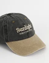 Gorra de gabardina color gris oscuro con visera color beige. Tiene bordado frontal con la palabra "Sunlight" y la frase "Follow your destiny".