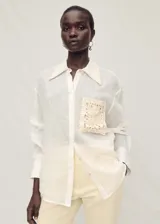 Camisa blanca de ramio con cuello camisero, cierre frontal con botones, bolsillo de parche con detalle bordado y mangas largas abotonadas.
