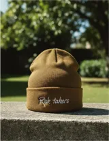 Gorro de lana sintética color marrón con bordado frontal de la frase "Risk takers" en color blanco.
