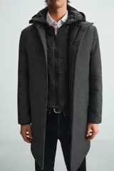 Abrigo gris oscuro con cuello doble y capucha desmontable con cremallera. Tiene manga larga, bolsillos en la cadera, bolsillo interior y abertura central en la espalda. El cierre frontal es con cremallera oculta por solapa con botones a presión.