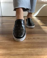 Zapatilla marrón de cuero con diseño entretejido, elástico lateral y suela blanca.