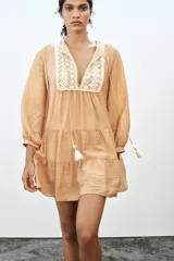 Vestido corto color beige con cuello redondo, cordón con borlas y manga larga. Presenta bordados combinados a contraste en la pechera, cierre frontal con botones ocultos, forro interior y bajo con volantes.