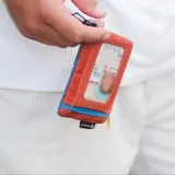 Billetera pequeña tipo pouch con múltiples compartimentos con cierre de cremallera en bloques de color: rojo, blanco, azul y gris oscuro. Incluye tiradores de cremallera amarillos y un mosquetón negro con cinta estampada con el logo de la marca.