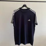 Remera Adidas Essentials color azul marino con cuello redondo y mangas cortas. Presenta las tres tiras blancas características de la marca en los hombros y el logo bordado en el pecho.