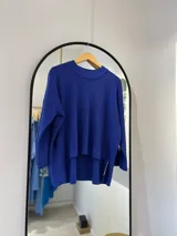 Sweater azul francia de punto con cuello a la base y manga pegada. Tiene hilado metalizado, delantera más corta que la parte trasera y aberturas en los laterales.