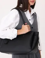 Cartera shopper de cuerina color negro con manijas superiores y charm a contratono. Tiene broche con botón para cerrar y cinturones laterales.