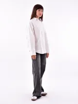 Camisa blanca con rayas verticales negras, de corte holgado, manga larga con puños abotonados y bolsillo en el pecho.