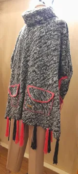 Poncho artesanal gris con cuello alto, bolsillos delanteros con borde rosa y flecos rosas y negros en el ruedo.