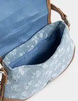 Bandolera de denim celeste con estampado floral blanco, detalles en cuero sintético marrón y correa de hombro ajustable. Cierre con hebilla metálica.