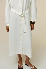 Vestido midi blanco de crepe con cuello redondo y escote pico, manga larga con puños y cinturón de la misma tela con hebilla metálica. Cierre frontal con botones metálicos.