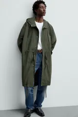 Parka verde militar de tejido técnico ligero, corte oversize, cuello alto con capucha ajustable, manga larga con puños elásticos, cintura ajustable con cordón interno, bolsillos con solapa, bajo asimétrico con abertura trasera ajustable y cierre frontal con cremallera oculta y botones a presión.