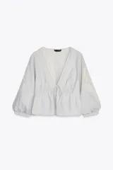 Blusa corta color celeste con textura arrugada, escote en V con puntilla, lazo ajustable en el frente y mangas 3/4 abullonadas con bordados florales a contraste.