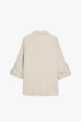 Camisa color marrón claro de cuello solapa y manga larga con trabilla y botón para remangar. Tiene un bolsillo de plastrón en el delantero, bajo asimétrico y cierre frontal con botones ocultos por solapa.