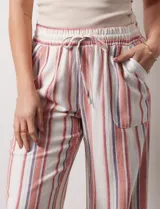 Pantalón de lino color blanco con rayas verticales rosas y celestes. Tiene pretina ancha elastizada con lazo de ajuste y bolsillos inclinados en la parte delantera.