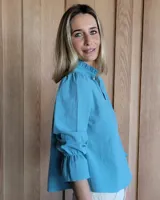 Blusa color beige con cuello con volados, mangas largas abullonadas con puños elásticos y cierre con botones en la parte delantera.