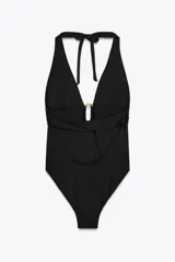 Traje de baño enterizo negro con escote halter atado al cuello y escote en V pronunciado. Presenta un aro metálico dorado en el centro del torso y espalda baja descubierta con banda cruzada.
