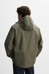 Parka verde militar confeccionada en tejido técnico ligero resistente al viento, con cuello alto, capucha, manga larga con puños ajustables, bolsillos de plastrón con solapa en la cadera, bolsillo interior, bajo ajustable con elásticos y cierre frontal con cremallera oculta por solapa con botones a presión.