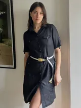 Vestido camisero negro de lino con cinturón de tela a rayas blancas y negras con hebilla dorada. Tiene cuello camisero, cierre frontal con botones, mangas cortas con trabillas y bolsillos en el pecho y laterales.