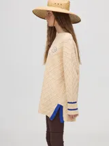 Sweater color crema con tejido en ochos, cuello redondo, puños acanalados con doble raya azul y tajos a los costados.