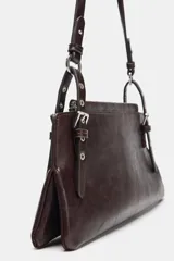 Bolso de hombro de formato alargado, color marrón oscuro con acabado ligeramente desgastado. El asa de hombro es delgada y ajustable mediante hebillas metálicas. Presenta detalles de tachas o remaches metálicos en los puntos de unión del asa.