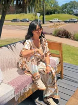 Pantalón ancho de satén con cintura elástica y estampado de motivos florales y cadenas en tonos marrones, beige y dorado sobre fondo blanco. Presenta un borde inferior con detalles en color turquesa.