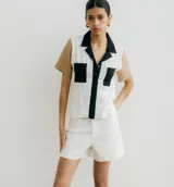 Camisa de manga corta color blanco con cuello negro, tapeta de botones negra y bolsillos cuadrados negros. Las mangas son color beige.