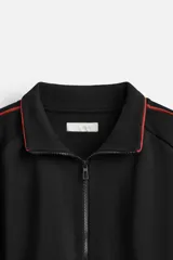 Chaqueta negra con detalles en rojo, cuello alto, cierre frontal con cremallera, bolsillos laterales y puños elásticos.