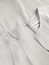 Blusa de satén color gris claro, con cuello redondo y abertura en forma de gota. Tiene mangas cortas con costuras en los hombros y cierre con broche en la espalda.