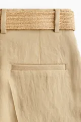 Short color beige de corte holgado con tiro alto, bolsillos laterales discretos, bolsillos atrás insertados y cierre con botón y corchete ocultos. Piernas con pinzas y pliegues adelante y atrás. Incluye cinturón ancho extraíble de rafia con hebilla cuadrada.