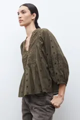 Blusa color verde oliva de manga 3/4 con volumen, cuello mao con abertura en V y botones. Presenta bordados y detalles de broderie calado en el canesú frontal y en las mangas. El cuerpo tiene un corte peplum con frunces.