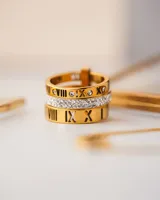 Anillo triple dorado de acero quirúrgico con baño de oro 18k. Dos de los anillos tienen números romanos grabados y el anillo central tiene circonitas blancas.