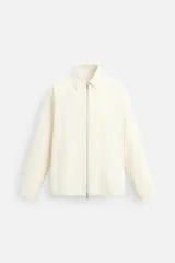 Sobrecamisa color crema de tejido técnico ligero, corte regular fit, cuello solapa, manga larga con puños abotonados, bajo ajustable con elástico en laterales y cierre frontal con cremallera.