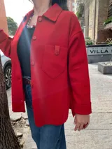 Camisa roja tejida en lana con cuello clásico, manga larga y cierre frontal con botones. Tiene un bolsillo de parche en el pecho con detalle de cuero.