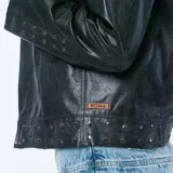 Chaqueta de cuero negra con cuello clásico, cierre frontal asimétrico con cremallera, solapas con muescas y detalles de tachas plateadas en los hombros y puños.