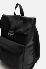 Mochila negra de tejido técnico con solapa y hebilla. Tiene un bolsillo frontal, un bolsillo interior con cierre y un compartimento para dispositivos electrónicos de hasta 13 pulgadas. La parte posterior es acolchada y tiene una banda de sujeción para equipaje.