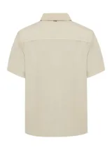 Camisa de manga corta color beige, con cuello clásico, cierre frontal con botones y bolsillo en el pecho izquierdo.