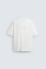 Camiseta blanca de corte holgado, cuello redondo y manga corta. Estampado frontal con texto en color negro y parche en la espalda. Detalle de pliegues en la espalda.