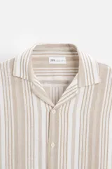 Camisa de manga corta con cuello bowling y corte relaxed fit. Diseño a rayas verticales en tonos beige y blanco. Cierre frontal con botones y bajo con aberturas laterales.