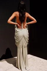 Vestido largo color beige topo, con escote halter y profundo escote pico pronunciado. Presenta un diseño drapeado con volumen en la zona del busto y espalda descubierta.