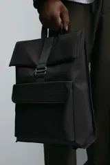 Mochila negra de tejido técnico con solapa y hebilla. Tiene un bolsillo frontal, un bolsillo interior con cierre y un compartimento para dispositivos electrónicos de hasta 13 pulgadas. La parte posterior es acolchada y tiene una banda de sujeción para equipaje.