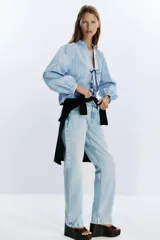Camisa celeste con rayas verticales marrones y blancas, cuello redondo con escote en pico y mangas largas abullonadas. Cierre frontal con lazos con pespuntes marcados.