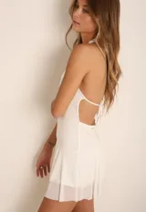 Minivestido blanco de corte ajustado, con cuello halter y falda con volados. Presenta un detalle de abertura en el pecho con aplique de strass.