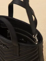 Cartera tote pequeña de cuero sintético negro con diseño acolchado en forma de espiga. Tiene doble asa corta superior, correa larga ajustable y desmontable, y logo metálico frontal con forma de labios.