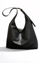 Cartera negra de cuero sintético con textura granulada, forma trapezoidal y asa de hombro ajustable con hebilla metálica.